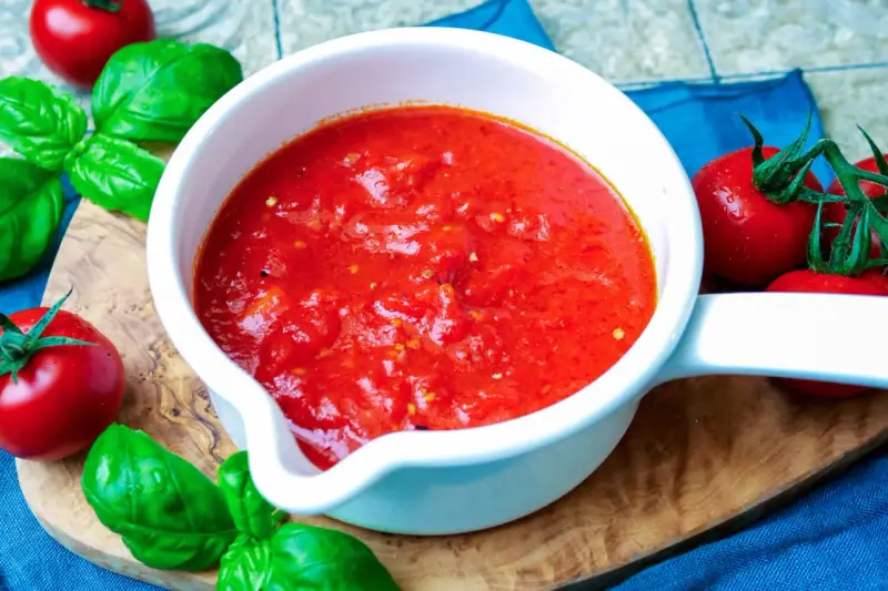 Frische Tomatensauce selber machen: Ein weißer Topf mit leuchtend roter Sauce, umgeben von reifen Tomaten und Basilikum auf einem Holzbrett.