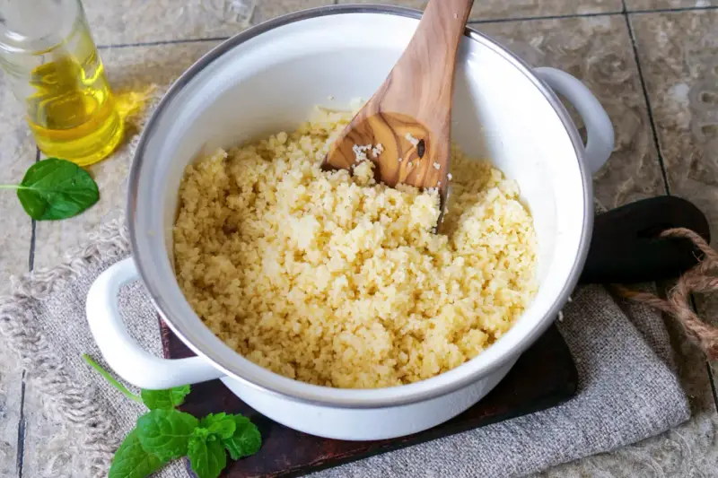 Ein Holzlöffel lockert den frisch zubereiteten Couscous in einem Topf auf. Daneben stehen Öl und Minze.